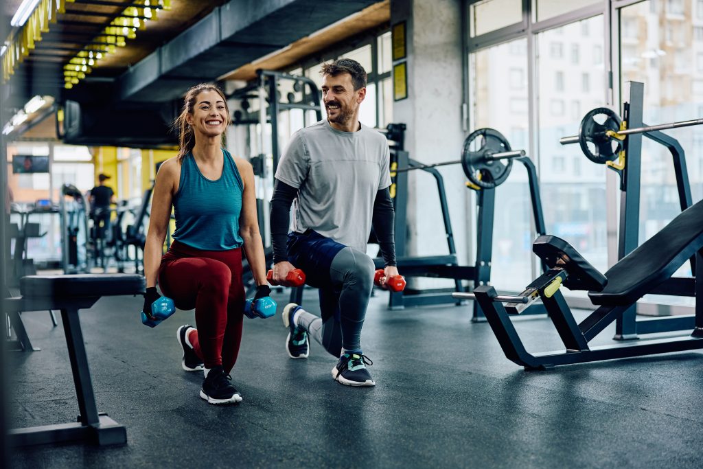 A woman and a man performing dumbbell lunges side-by-side in a gym. Both are smiling and appear to be enjoying the workout. The gym has weightlifting equipment, benches, and large windows in the background.