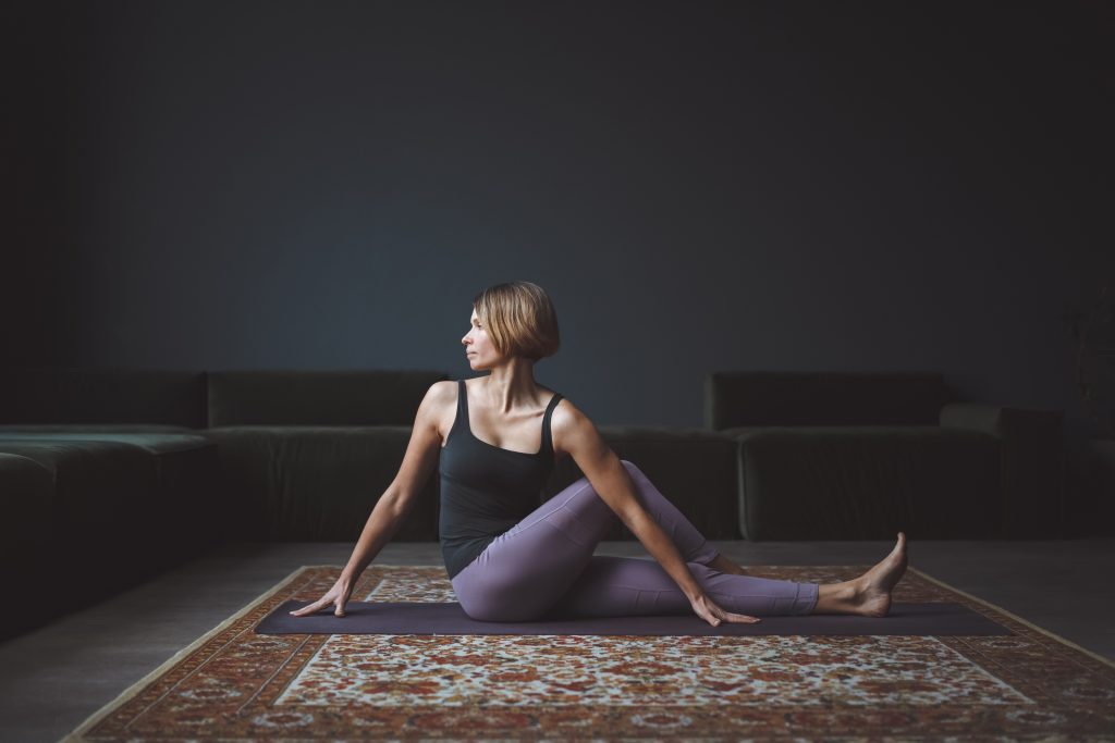 Person seated on a yoga mat performing a seated spinal twist stretch, with one leg bent and the other extended, in a calm living room setting with a sofa and rug in the background.
