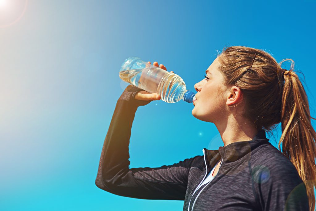 Woman drinking water from a plastic bottle outdoors under a clear blue sky, wearing athletic clothing and emphasizing hydration during physical activity.