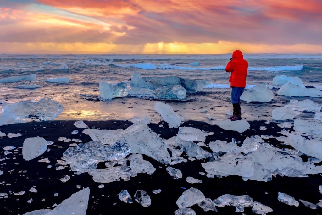 **Alt text:**
Person in a red jacket standing on an icy beach surrounded by chunks of ice, looking out over the ocean at sunrise or sunset under a colorful sky.
