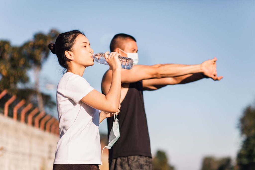 **Alt text:**
Two people outdoors during a workout break, with one person drinking water from a bottle while the other stretches their arms, emphasizing hydration and recovery during exercise.
