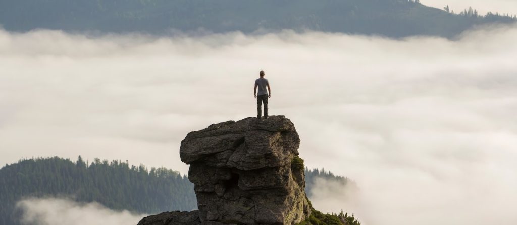**Alt text:**
Person standing on top of a rocky cliff overlooking a fog-filled valley with forested hills in the distance, symbolizing high altitude and outdoor adventure.