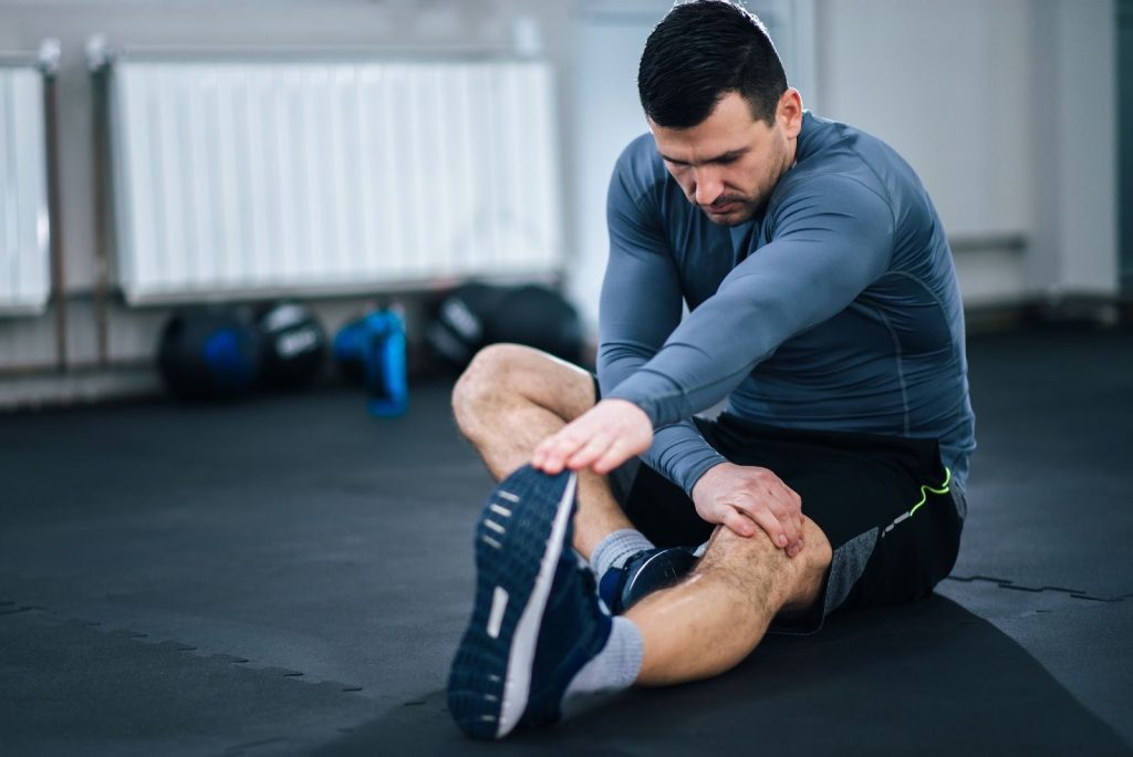 **Alt text:**
Man seated on the floor performing a seated hamstring stretch, reaching toward his extended leg in a gym setting with exercise equipment in the background.