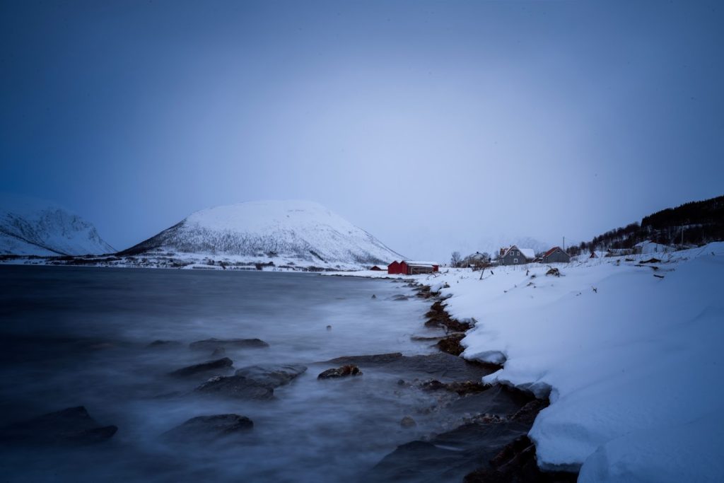 Snow-covered coastal landscape with icy shoreline, calm water, and distant mountains under an overcast sky, with a few small buildings visible along the shore.