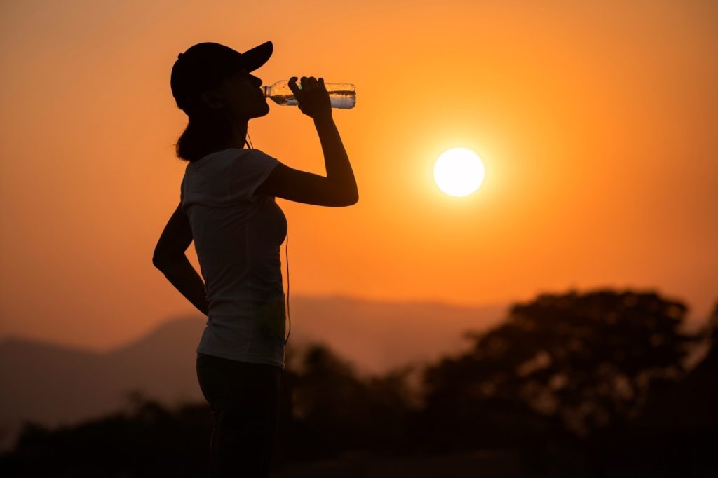 Silhouette of a person drinking water from a bottle at sunset, standing outdoors against an orange sky with the sun low on the horizon, emphasizing hydration during outdoor activity.