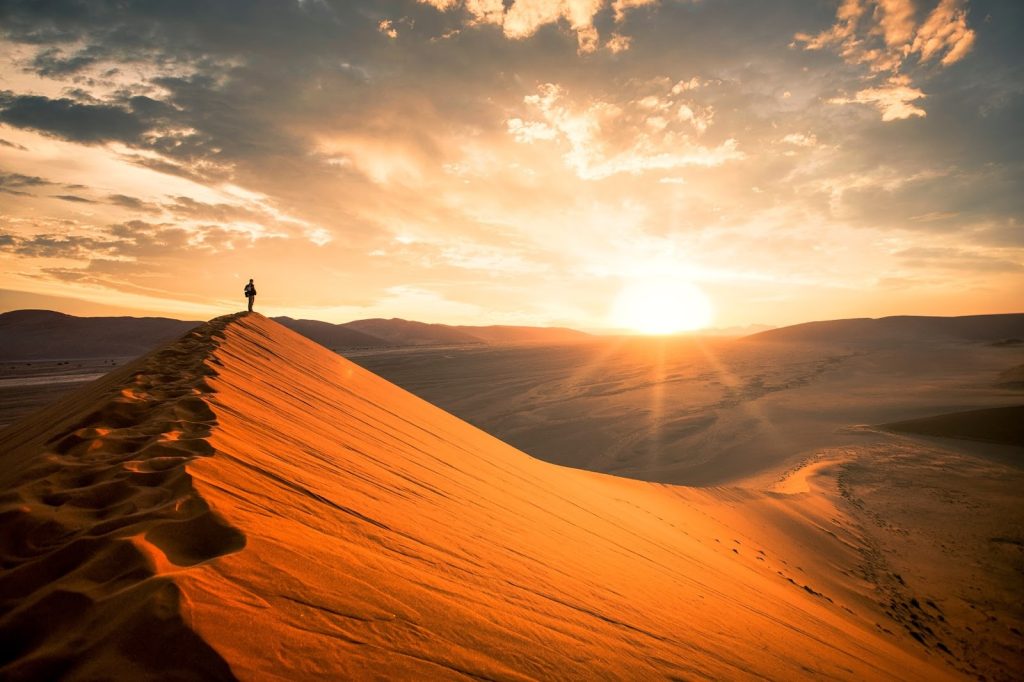 **Alt text:**
Person standing on the crest of a sand dune at sunset, with sweeping desert landscape and warm golden light highlighting the dune’s curves, symbolizing heat and environmental exposure.