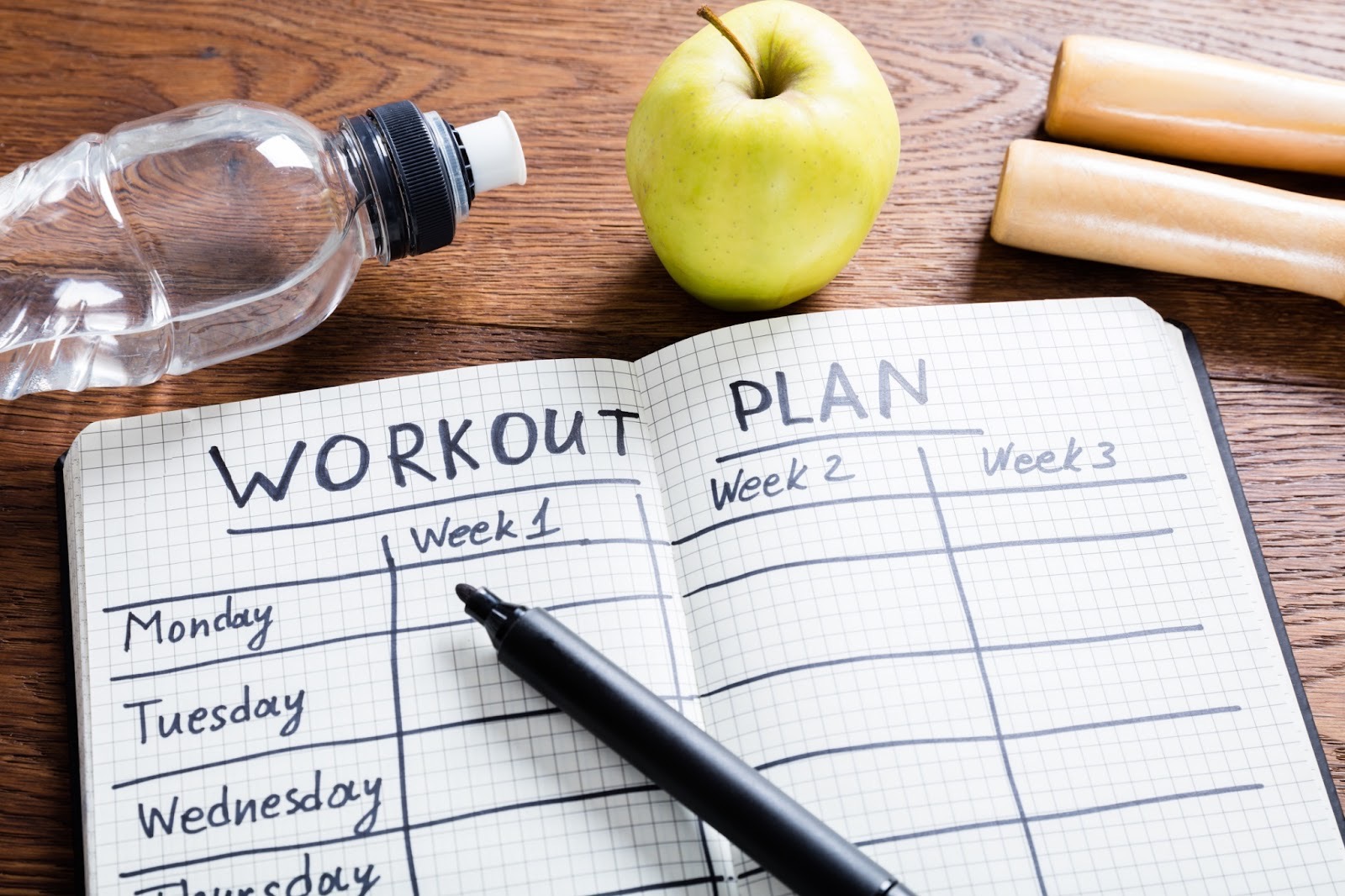 Open notebook showing weekly workout plan schedule with pen, apple, water bottle, and fitness bars on wooden table.