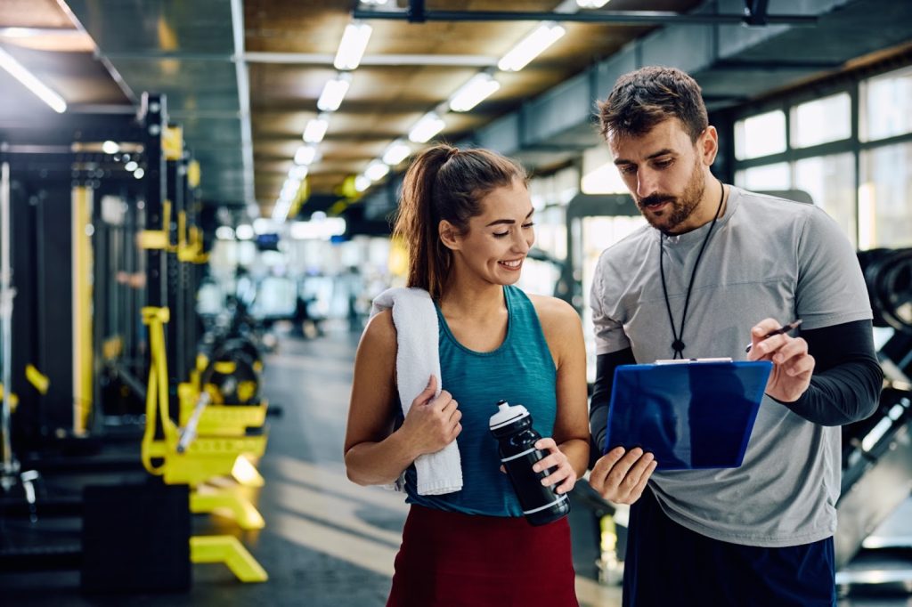 Personal trainer reviewing workout plan on clipboard with smiling gym client holding water bottle in a modern fitness center.