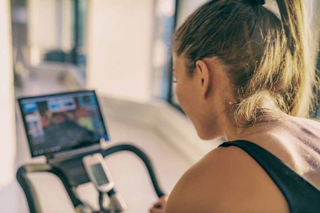 A woman with blonde hair viewed from behind, riding a stationary bike while watching a screen mounted on the handlebars.
