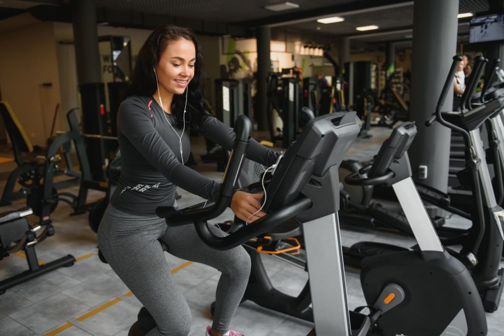 A woman in gray athletic wear using an exercise bike in a dimly lit gym with equipment visible in the background.