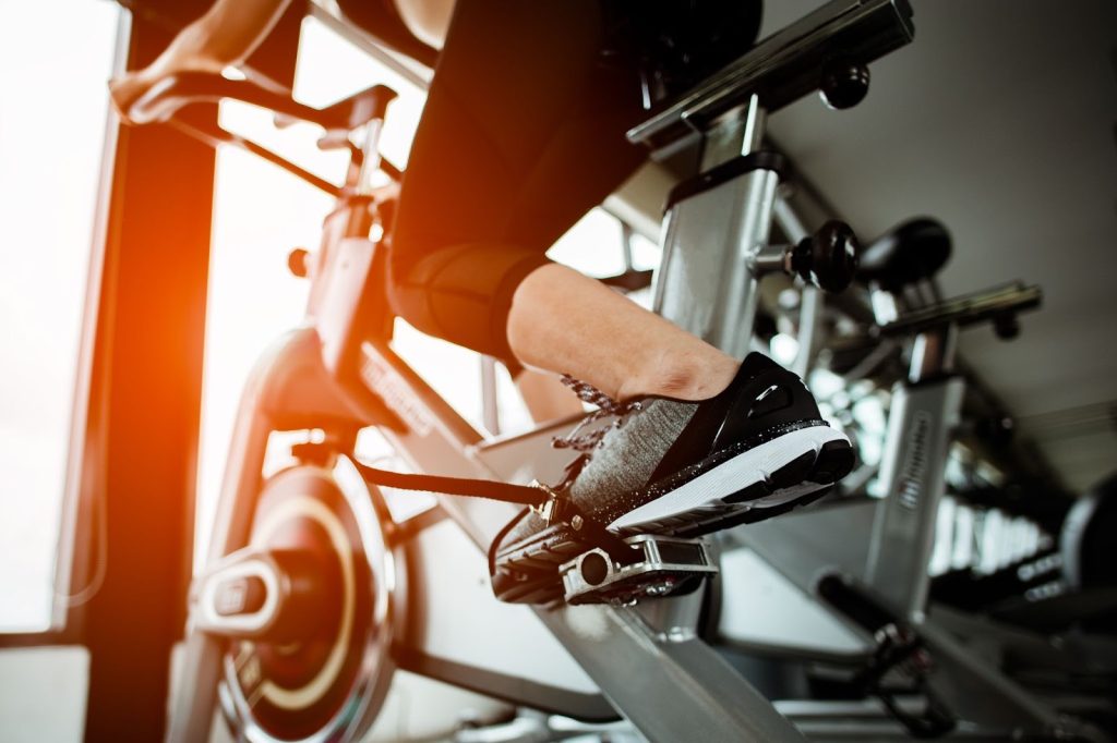 A close-up of a person's feet clipped into the pedals of a stationary bike, with warm backlit gym lighting in the background.