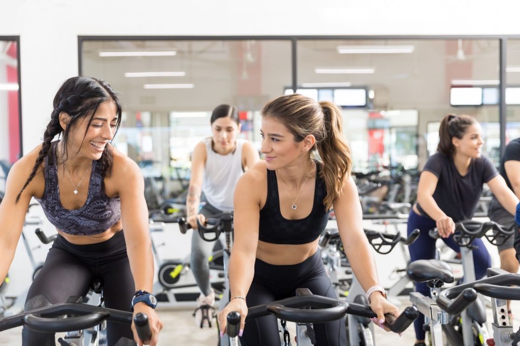 Three women smiling and riding stationary bikes together in a bright spin class at a gym.