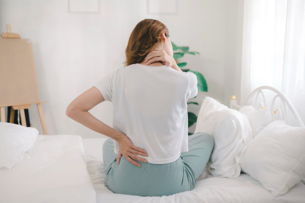 A woman in a white top and light blue pants sitting on a white bed and reaching to hold her lower back