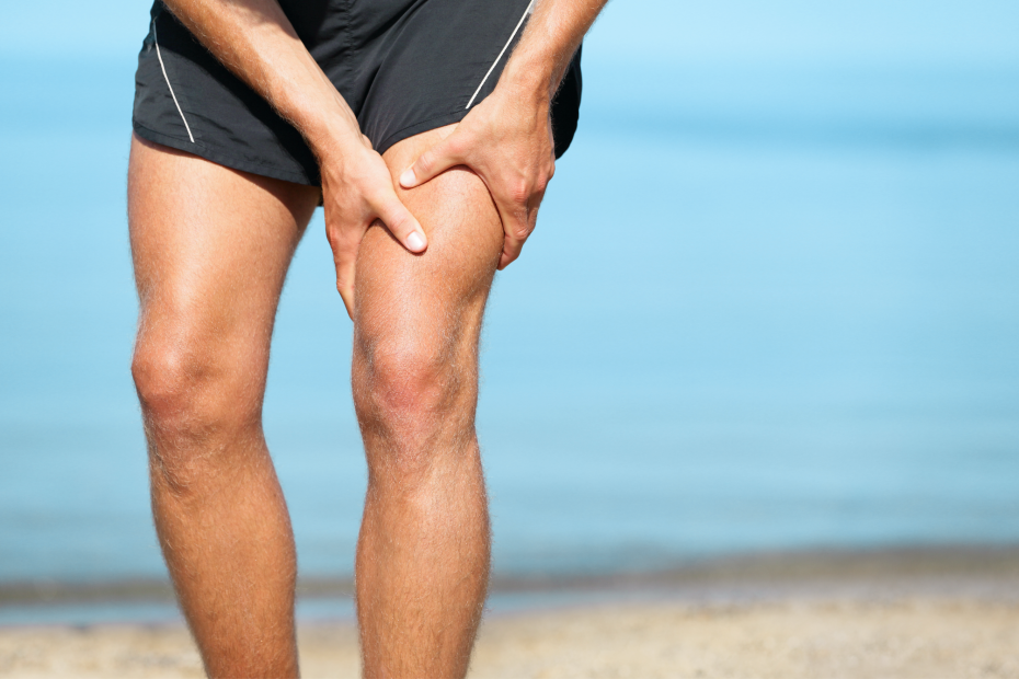 Close-up of a person in black shorts gripping their cramped leg with both hands against a blurred ocean background,