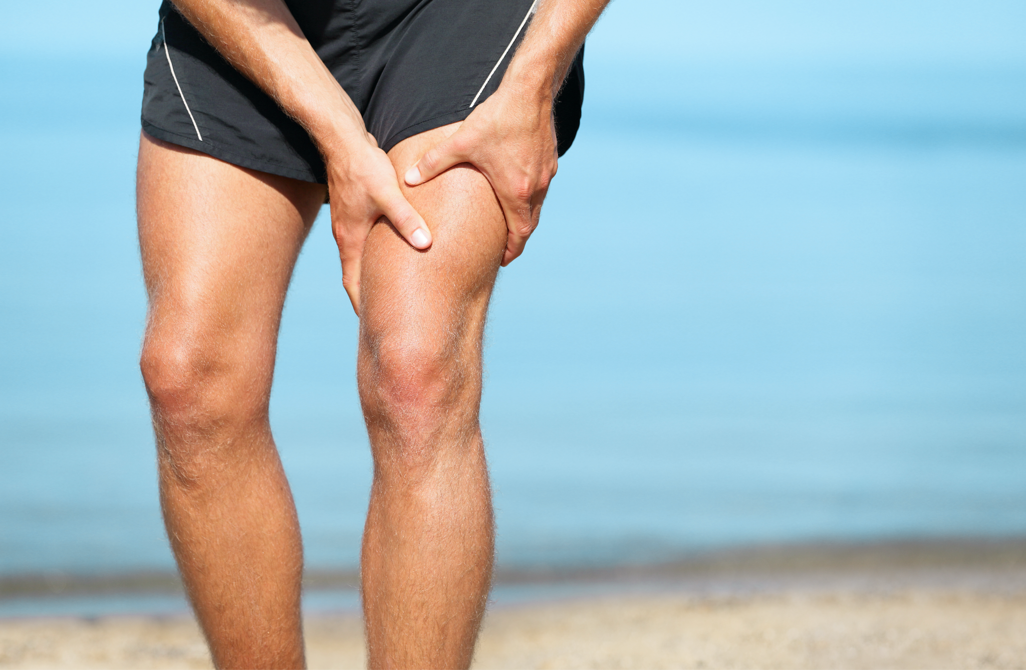Close-up of a person in black shorts gripping their cramped leg with both hands against a blurred ocean background,