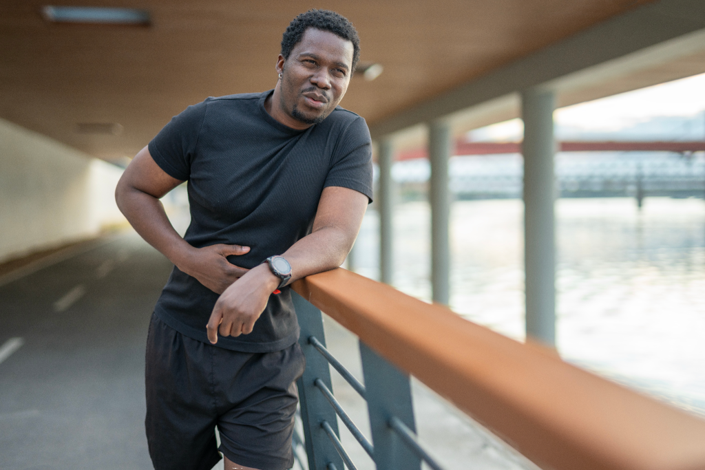 A Black man in a dark t-shirt and joggers resting while holding his stomach because of a muscle cramp
